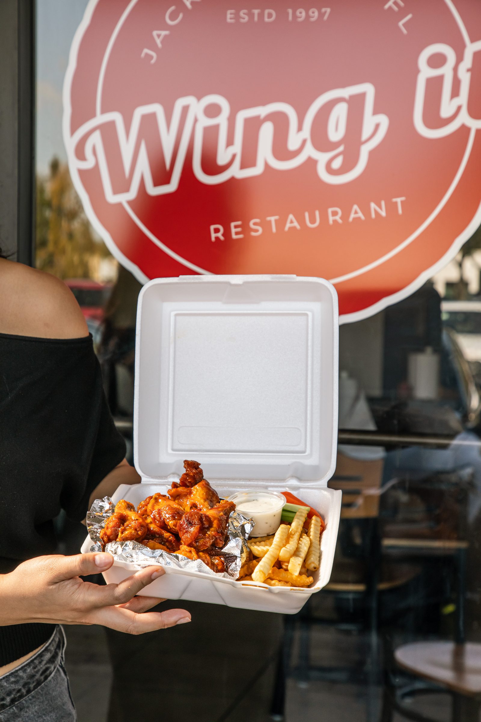 A takeout box of Wing It wings held up outside the restaurant